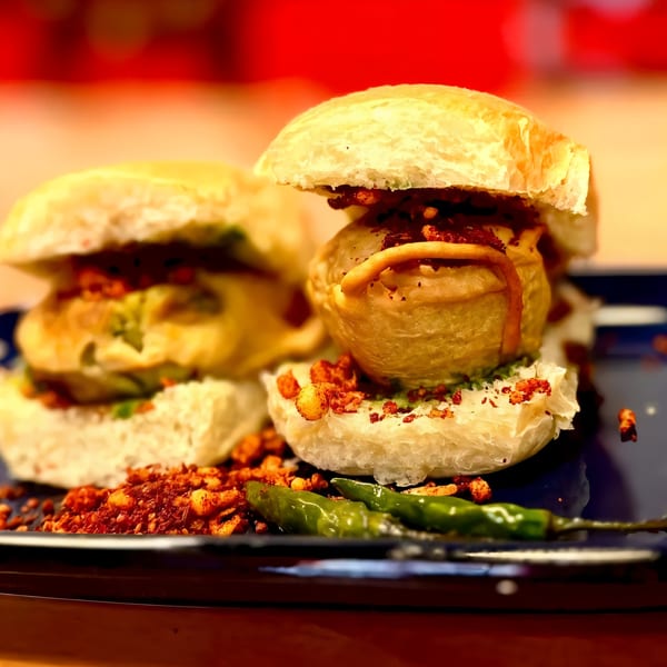 Two Indian vada pavs with green chilies and spicy red chutney on a dark plate.