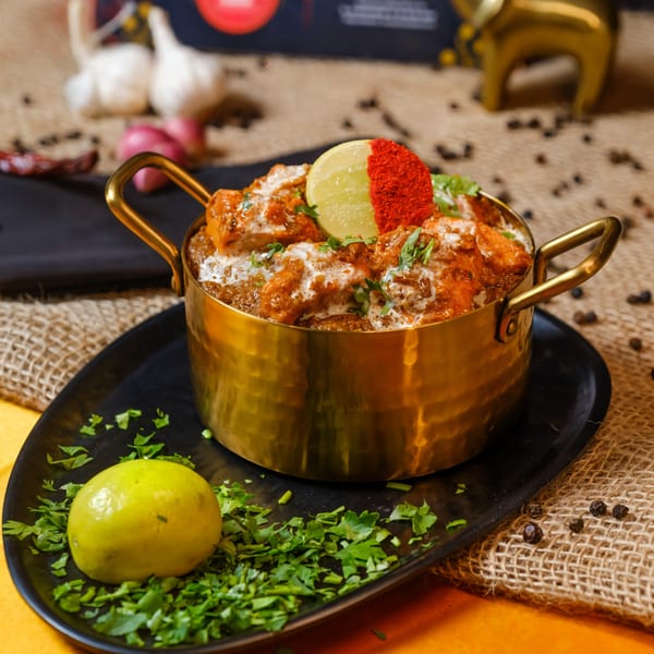 Indian butter chicken in a brass pot, garnished with cream, cilantro, and a lemon slice.