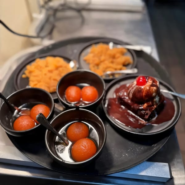 A black tray with four bowls of Gulab Jamun, two plates of orange Indian dessert, and a chocolate brownie.